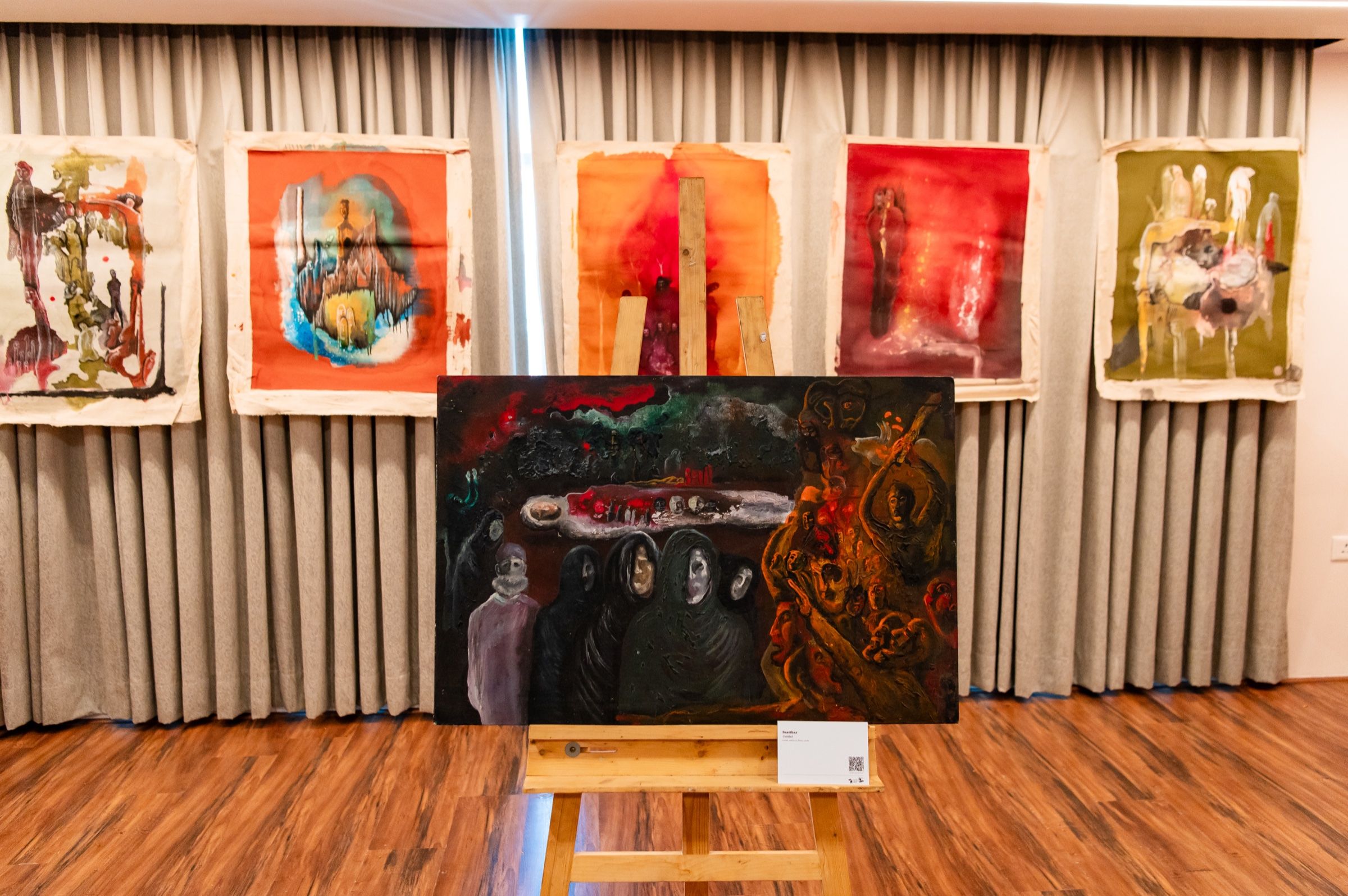 A dark figurative painting on an easel framed by colourful paintings hung on fabric.
